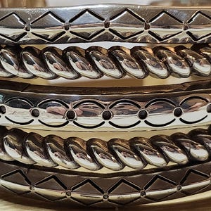 K&ouml;nnte beinhalten: Nahaufnahme mehrerer silberfarbener Armreifen. Die Armreifen weisen verschiedene dekorative Muster auf, darunter Rautenformen, gedrehtes Seil und ovale Aussparungen. Der Schmuck ist gestapelt und zeigt die verschiedenen Designs und Texturen.