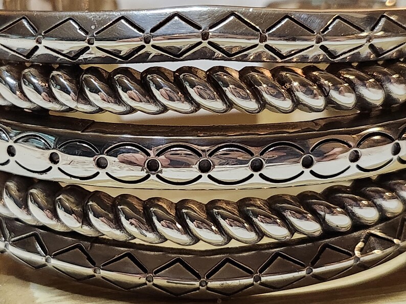 K&ouml;nnte beinhalten: Nahaufnahme mehrerer silberfarbener Armreifen. Die Armreifen weisen verschiedene dekorative Muster auf, darunter Rautenformen, gedrehtes Seil und ovale Aussparungen. Der Schmuck ist gestapelt und zeigt die verschiedenen Designs und Texturen.