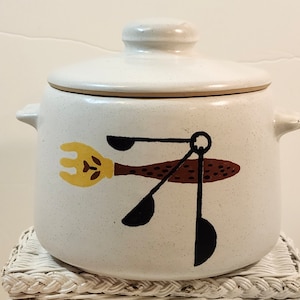May include: A white ceramic kitchen canister with a lid, featuring a brown and yellow fork and spoon design. The canister has two side handles and a speckled finish, perfect for storing kitchen essentials.