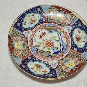 May include: A round, decorative ceramic plate with a gold rim. The plate features a central floral design surrounded by intricate patterns in red, blue, and gold. The plate's diameter is approximately 6 inches.