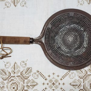 May include: An antique, round metal trivet with a perforated surface and a wooden handle. The trivet is a rusty brown color, with a wooden handle and a leather loop for hanging. The trivet is designed to protect surfaces from heat.