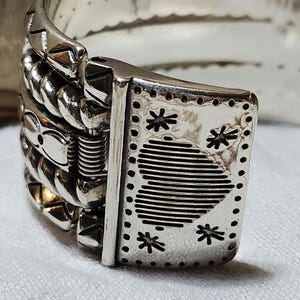 K&ouml;nnte beinhalten: Nahaufnahme eines silberfarbenen Armbandverschlusses. Der Verschluss hat eine rechteckige Platte mit sternf&ouml;rmigen Aussparungen und einem gestreiften Design. Die Kette des Armbands hat ein strukturiertes, geflochtenes Aussehen, mit einem sichtbaren Federmechanismus.