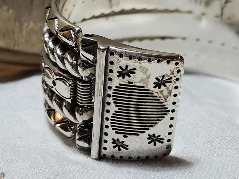 K&ouml;nnte beinhalten: Nahaufnahme eines silberfarbenen Armbandverschlusses. Der Verschluss hat eine rechteckige Platte mit sternf&ouml;rmigen Aussparungen und einem gestreiften Design. Die Kette des Armbands hat ein strukturiertes, geflochtenes Aussehen, mit einem sichtbaren Federmechanismus.