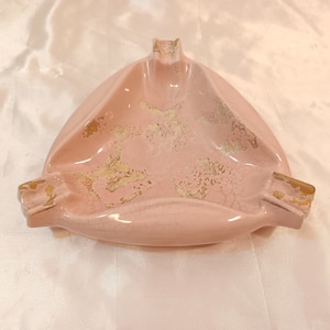 May include: A pink ceramic ashtray with gold accents. The ashtray is shaped like a triangle with three rounded sides. The gold accents are in a crackled pattern.