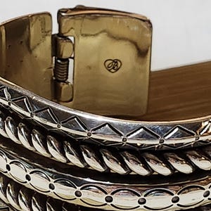 K&ouml;nnte beinhalten: Nahaufnahme eines silberfarbenen Armreifs mit aufwendigen Details. Das Armband hat einen Klappverschluss mit goldfarbenem Finish und einem kleinen herzf&ouml;rmigen Emblem. Das Design umfasst geometrische und seilartige Muster.