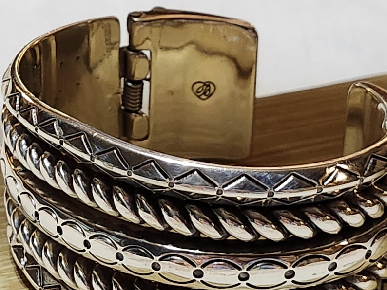 K&ouml;nnte beinhalten: Nahaufnahme eines silberfarbenen Armreifs mit aufwendigen Details. Das Armband hat einen Klappverschluss mit goldfarbenem Finish und einem kleinen herzf&ouml;rmigen Emblem. Das Design umfasst geometrische und seilartige Muster.