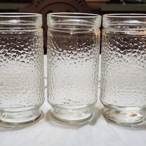 May include: Three clear glass tumblers with a textured, pebbled surface. The glasses have a cylindrical shape with a slightly flared base and a smooth rim. They are arranged side by side on a white surface.