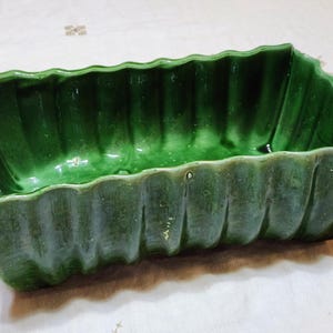 May include: A rectangular ceramic planter with a glossy, deep green glaze. The exterior and interior walls feature a consistent vertical fluted or ribbed texture, creating a scalloped edge along the top rim of the vessel.