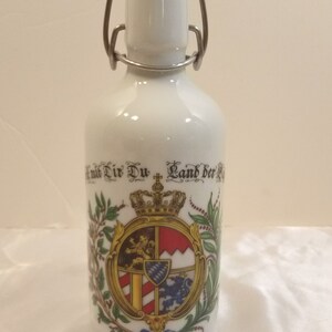 May include: White ceramic bottle with a silver hinged lid and a colorful coat of arms featuring a crown, lion, and other heraldic symbols. The bottle has the text "Mit Dir Du Land der Bayern" printed on it.