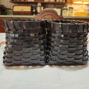 May include: Two dark brown woven baskets. The baskets are cylindrical with a flared base and a slightly wider top. The weave pattern is horizontal, creating a textured surface. The baskets are sitting on a floral patterned tablecloth.