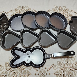 May include: A collection of metal baking pans in various shapes, including a Pinocchio, snowman, heart, bunny, and round tart pans. Each pan has a dark gray finish and a black handle. The pans are arranged on a patterned white surface.