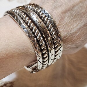 K&ouml;nnte beinhalten: Eine Sammlung silberfarbener Metallarmb&auml;nder mit verschiedenen gepr&auml;gten Mustern, darunter Diamanten, Seile und florale Designs. Die Armb&auml;nder sind gestapelt, um ihre komplizierten Details und Texturen hervorzuheben. Der Schmuck wird am Handgelenk getragen.