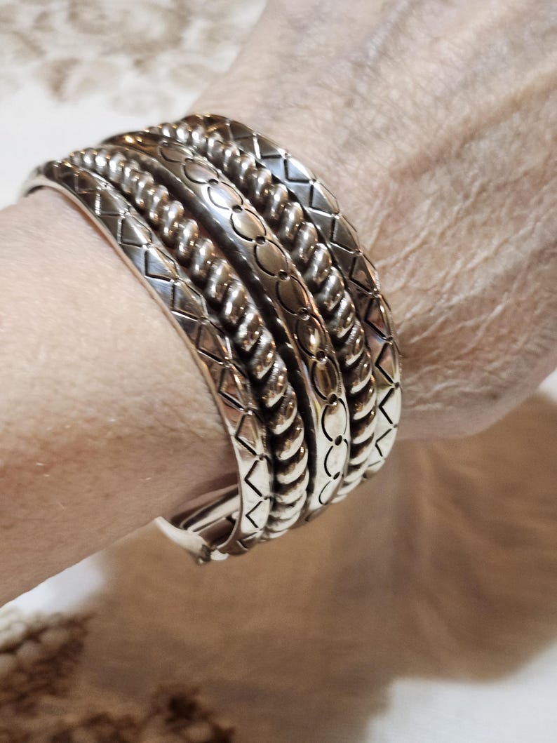 K&ouml;nnte beinhalten: Eine Sammlung silberfarbener Metallarmb&auml;nder mit verschiedenen gepr&auml;gten Mustern, darunter Diamanten, Seile und florale Designs. Die Armb&auml;nder sind gestapelt, um ihre komplizierten Details und Texturen hervorzuheben. Der Schmuck wird am Handgelenk getragen.