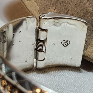 K&ouml;nnte beinhalten: Nahaufnahme eines silberfarbenen, klappbaren Armbandverschlusses. Der Verschluss ist rechteckig mit einem kleinen herzf&ouml;rmigen Logo. Das Armband hat ein strukturiertes, gemustertes Design. Der Scharniermechanismus enth&auml;lt eine sichtbare Feder.