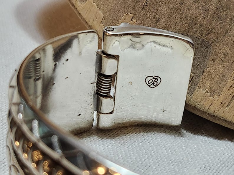 K&ouml;nnte beinhalten: Nahaufnahme eines silberfarbenen, klappbaren Armbandverschlusses. Der Verschluss ist rechteckig mit einem kleinen herzf&ouml;rmigen Logo. Das Armband hat ein strukturiertes, gemustertes Design. Der Scharniermechanismus enth&auml;lt eine sichtbare Feder.