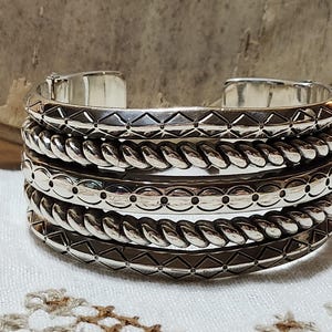 K&ouml;nnte beinhalten: Ein Stapel silberfarbener Metallarmreifen. Die Armreifen weisen verschiedene Texturen auf, darunter Rautenmuster, seilartige Drehungen und gepunktete Designs. Die Armreifen werden auf einer wei&szlig;en Oberfl&auml;che pr&auml;sentiert.