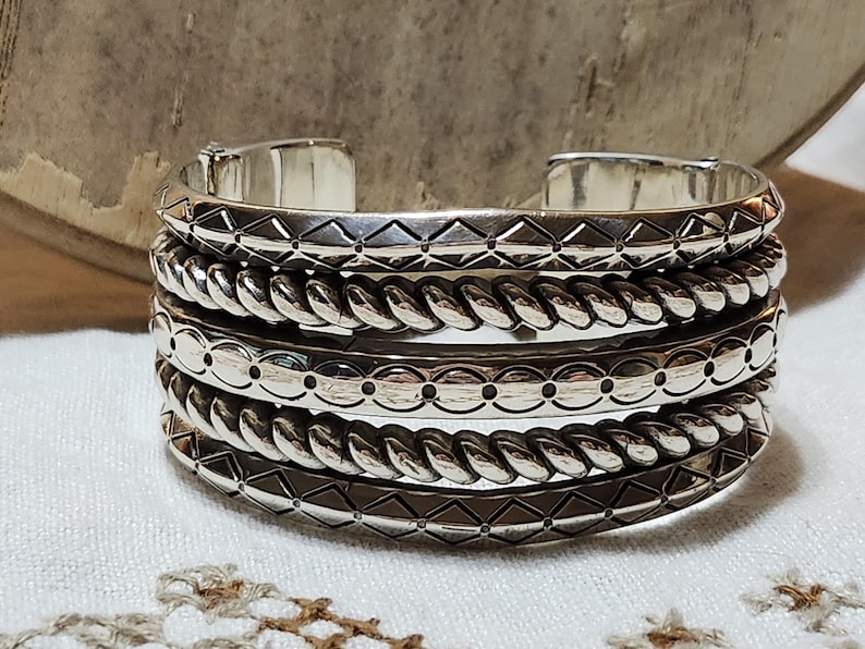 K&ouml;nnte beinhalten: Ein Stapel silberfarbener Metallarmreifen. Die Armreifen weisen verschiedene Texturen auf, darunter Rautenmuster, seilartige Drehungen und gepunktete Designs. Die Armreifen werden auf einer wei&szlig;en Oberfl&auml;che pr&auml;sentiert.