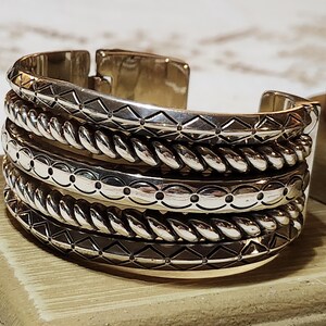 K&ouml;nnte beinhalten: Ein silbernes Armreif-Armband mit vier gestapelten B&auml;ndern. Die B&auml;nder weisen detaillierte Muster auf, darunter geometrische Formen, seilartige Texturen und gepunktete Designs. Das Armband wird auf einer Holzoberfl&auml;che pr&auml;sentiert.