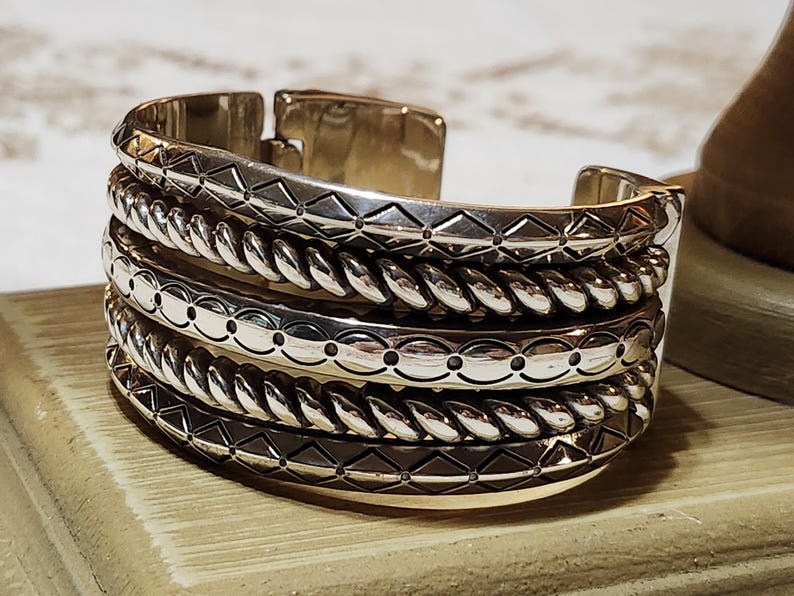 K&ouml;nnte beinhalten: Ein silbernes Armreif-Armband mit vier gestapelten B&auml;ndern. Die B&auml;nder weisen detaillierte Muster auf, darunter geometrische Formen, seilartige Texturen und gepunktete Designs. Das Armband wird auf einer Holzoberfl&auml;che pr&auml;sentiert.
