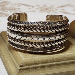 K&ouml;nnte beinhalten: Ein silbernes Armreif-Armband mit vier gestapelten B&auml;ndern. Jedes Band hat ein anderes Design: geometrisch, geflochten, gepunktet und geometrisch. Das Armband wird auf einer Holzoberfl&auml;che pr&auml;sentiert.