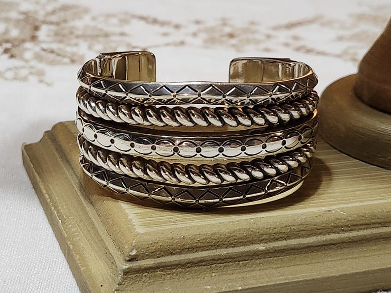K&ouml;nnte beinhalten: Ein silbernes Armreif-Armband mit vier gestapelten B&auml;ndern. Jedes Band hat ein anderes Design: geometrisch, geflochten, gepunktet und geometrisch. Das Armband wird auf einer Holzoberfl&auml;che pr&auml;sentiert.