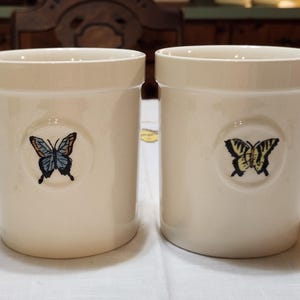 May include: Two off-white ceramic kitchen utensil holders. Each cylindrical holder features a butterfly design in a circular emblem. One butterfly is blue and black, the other is yellow and black.