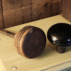 May include: Two door knobs are shown. One is a round, dark brown wooden knob with a metal spindle attached to its side. The other is a smooth, glossy, round black knob with a short, dark cylindrical base extending from its center.