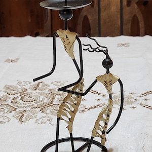 May include: A black metal candle holder featuring two stylized figures. The figures have black spherical heads and are adorned with beige woven material. The candle holder has a circular base and a small, round platform for a candle.