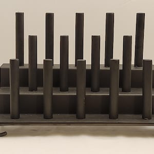 May include: A dark gray metal rack with two tiers and vertical cylindrical supports. The rack has a rectangular base with curved feet. The supports are evenly spaced and rise from the tiers, creating a structured design.