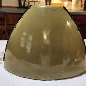 May include: A large, olive-green ceramic lampshade with a rounded, bell-like shape. The shade has a glossy finish and is trimmed with black metal accents along the top edge. The lampshade sits on a white tablecloth.