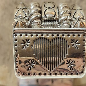 K&ouml;nnte beinhalten: Eine silberfarbene dekorative Metallbox mit herzf&ouml;rmigem Design und Schneeflocken-Akzenten. Die Oberseite zeigt stilisierte B&auml;ume und gedrehte, seilartige Elemente. Die Box hat eine rechteckige Form mit detaillierter, strukturierter Oberfl&auml;che.