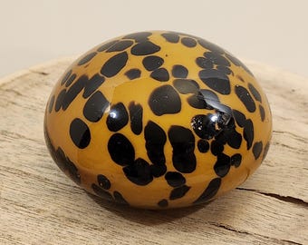 May include: A rounded, decorative glass object with a glossy finish. The object is amber-colored with irregular black spots, resembling a tortoise shell pattern. It sits on a wooden surface.
