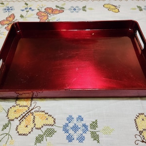 May include: A rectangular, red serving tray with built-in handles. The tray has a metallic finish and is designed for carrying items. The tray is approximately 16 inches long and 10 inches wide.