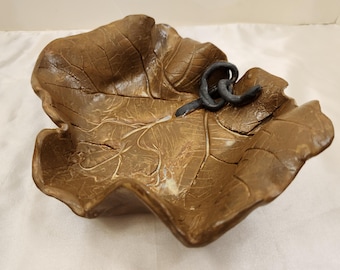 May include: A brown ceramic leaf-shaped bowl with a black metal accent. The bowl is textured to resemble a leaf.