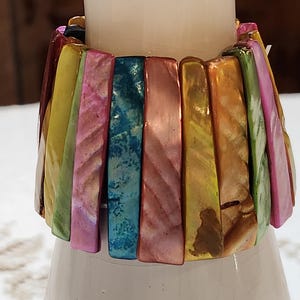 May include: A colorful bracelet with rectangular, iridescent beads in shades of pink, blue, yellow, and orange. The bracelet is wrapped around a white cylindrical object, creating a vibrant display of color and texture.