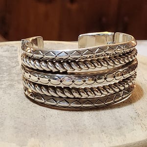 K&ouml;nnte beinhalten: Silbernes Armreif-Armband mit aufwendigen Details. Das Armband hat abwechselnde B&auml;nder mit geometrischen Mustern, seilartigen Texturen und gepunkteten Designs. Der Armreif wird auf einer hellen, strukturierten Oberfl&auml;che pr&auml;sentiert.