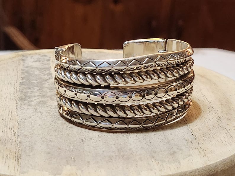 K&ouml;nnte beinhalten: Silbernes Armreif-Armband mit aufwendigen Details. Das Armband hat abwechselnde B&auml;nder mit geometrischen Mustern, seilartigen Texturen und gepunkteten Designs. Der Armreif wird auf einer hellen, strukturierten Oberfl&auml;che pr&auml;sentiert.