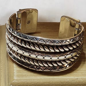 K&ouml;nnte beinhalten: Silberfarbenes Armreif-Armband mit aufwendigen Details. Das Armband hat mehrere B&auml;nder mit strukturierten Mustern, darunter seilartige Designs und geometrische Formen. Der Verschluss ist sichtbar, und das Armband scheint aus Metall zu sein.