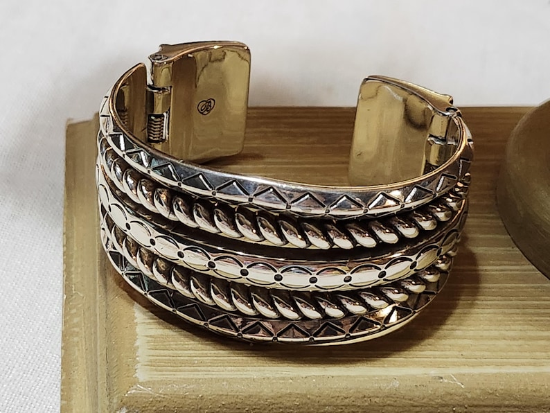 K&ouml;nnte beinhalten: Silberfarbenes Armreif-Armband mit aufwendigen Details. Das Armband hat mehrere B&auml;nder mit strukturierten Mustern, darunter seilartige Designs und geometrische Formen. Der Verschluss ist sichtbar, und das Armband scheint aus Metall zu sein.