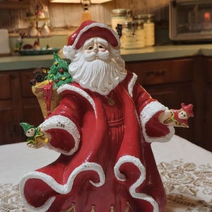 May include: A Santa Claus figurine wearing a long red coat with white fur trim and gold accents. Santa has a white beard, a red hat, and carries a sack filled with a green Christmas tree and gifts. Small elf figures are held in each outstretched hand.