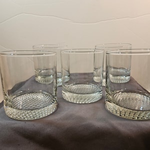 May include: Six clear glass tumblers with a textured bottom. The glasses are empty and arranged in a row on a gray surface.