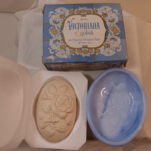 May include: A white soap dish with a raised floral design, a blue soap dish with a raised floral design, and a blue box with gold trim that reads "Avon Victorian Soap Dish and 'Special Occasion' Soap Net Wt 5 oz."