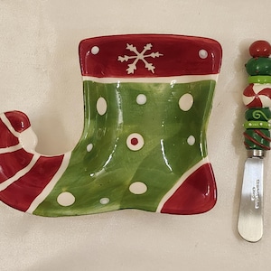 May include: A green and red ceramic Christmas stocking shaped dish with white polka dots and a snowflake design. It comes with a matching green and red candy cane-themed butter knife.