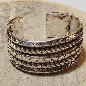 K&ouml;nnte beinhalten: Silberfarbenes Armreif-Armband mit aufwendigen Details. Das Armband hat mehrere strukturierte B&auml;nder, darunter Seil- und geometrische Muster. Der Klappverschluss ist ge&ouml;ffnet und zeigt den inneren Mechanismus. Ein stilvolles Schmuckst&uuml;ck.