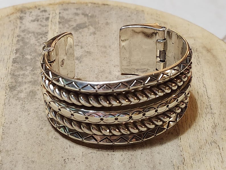 K&ouml;nnte beinhalten: Silberfarbenes Armreif-Armband mit aufwendigen Details. Das Armband hat mehrere strukturierte B&auml;nder, darunter Seil- und geometrische Muster. Der Klappverschluss ist ge&ouml;ffnet und zeigt den inneren Mechanismus. Ein stilvolles Schmuckst&uuml;ck.