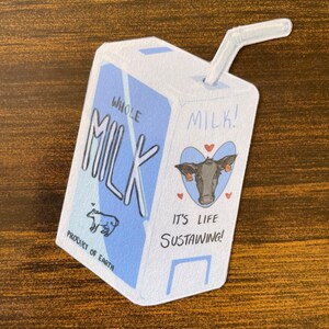 May include: A blue and white cartoon illustration of a milk carton with a red heart and a cow's face. The carton says "Whole Milk!" and "It's Life Sustaining!"  The carton also says "Product of Earth" and has a small cartoon cow on it.