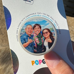 May include: A PopSockets phone grip and stand in its original white packaging with holographic accents. The product features a circular image of three people smiling, with text reading PopSockets, Phone Grip & Stand with Swappable Top.