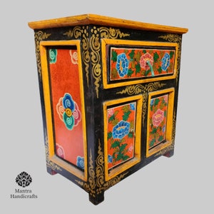 May include: A small black wooden cabinet with a single drawer and two doors. The cabinet is painted with colourful floral designs in shades of red, blue, green, and yellow. The cabinet has gold accents and a decorative border.