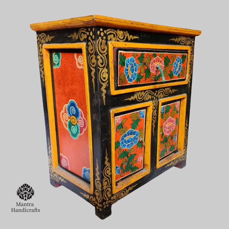 May include: A small black wooden cabinet with a single drawer and two doors. The cabinet is painted with colourful floral designs in shades of red, blue, green, and yellow. The cabinet has gold accents and a decorative border.