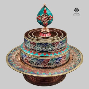 May include: A multi-tiered brass offering bowl with intricate designs and colourful turquoise and red stones. The bowl is topped with a finial featuring a turquoise teardrop shape and red stones. The bowl is on a base with a similar design.